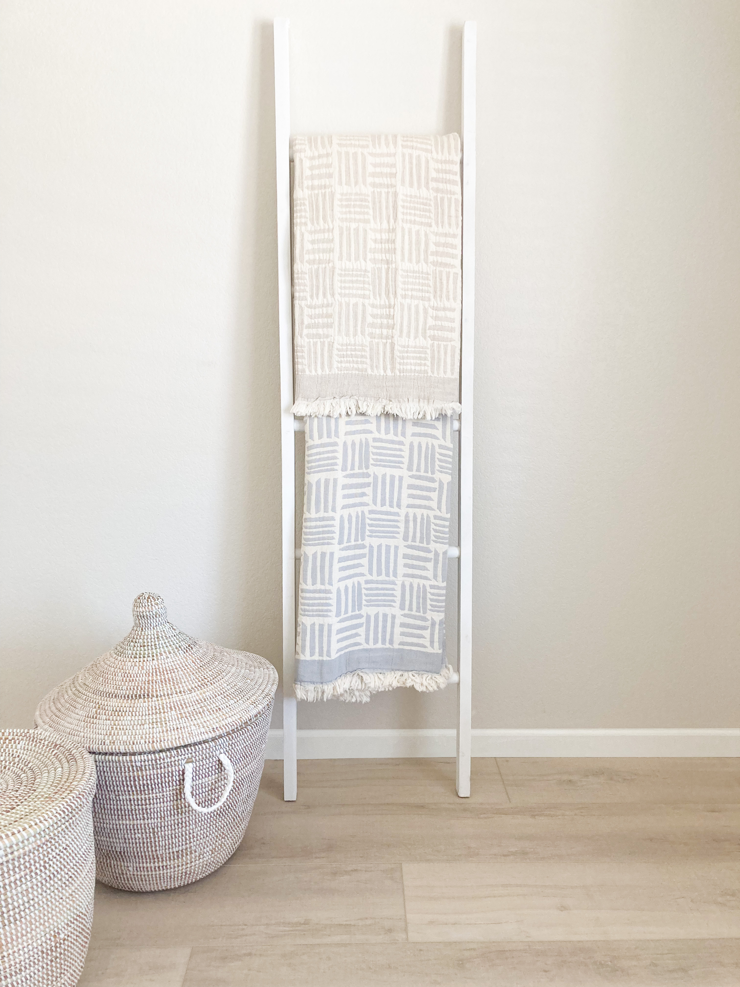2 blankets hanging on a blanket ladder. There top blanket is beige and ivory. The bottom blanket is light gray and ivory. Both blankets have a modern geometric pattern. There are 2 baskets with lids to the bottom left of the ladder.