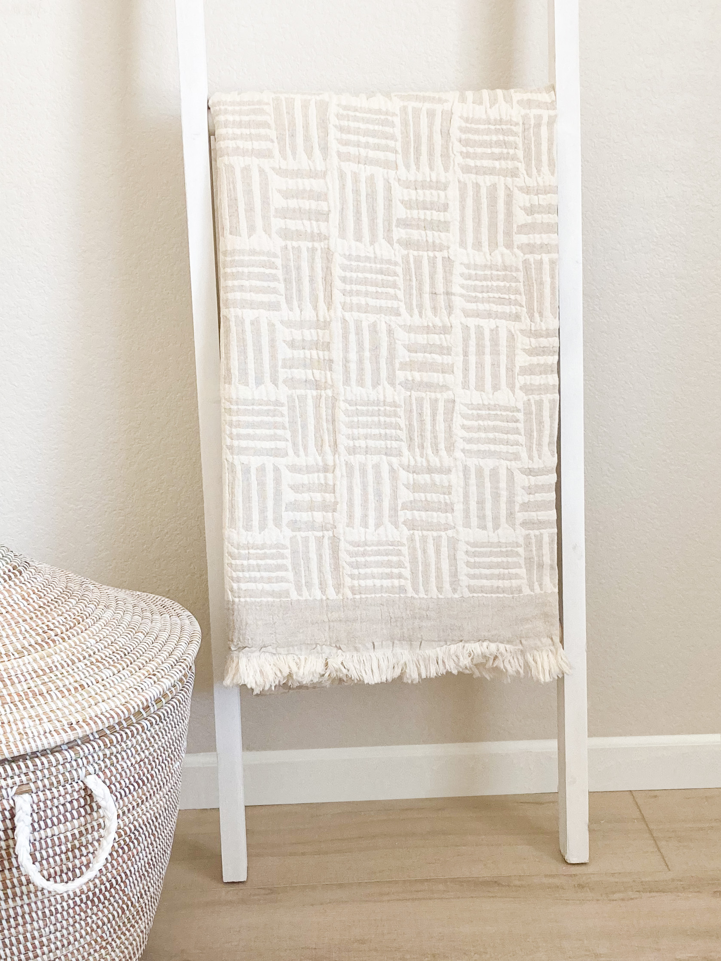 ivory and beige blanket hanging on a ladder. The blanket has a modern geometric pattern. There is a large basket with the lid to the bottom left of the blanket ladder.