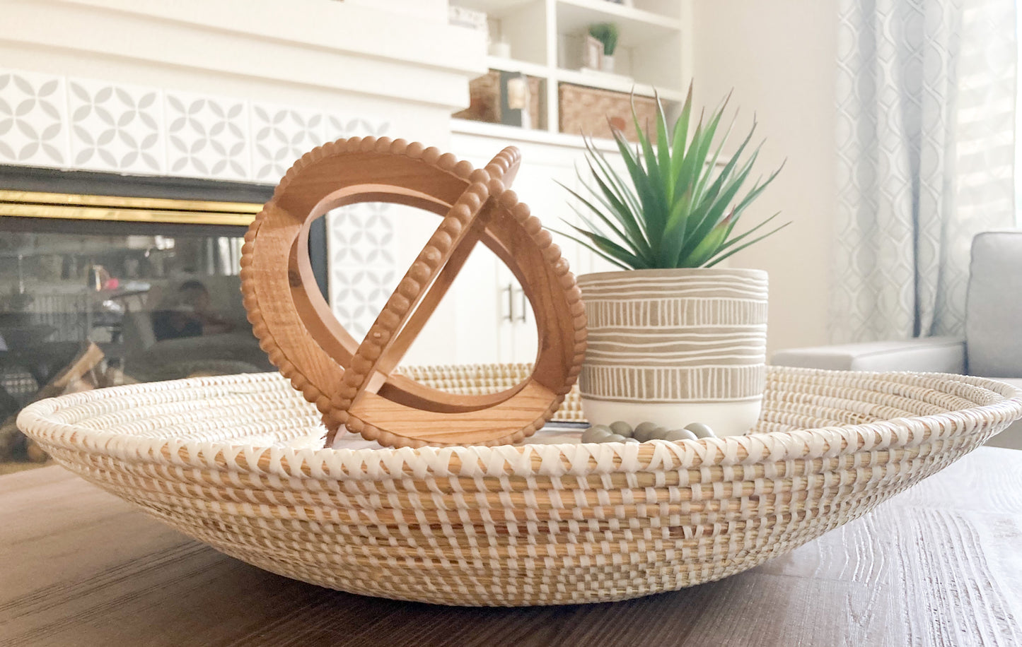 large tabletop round basket with group of decor items: wooden orb, faux plant, and beads- this is seen from the side view