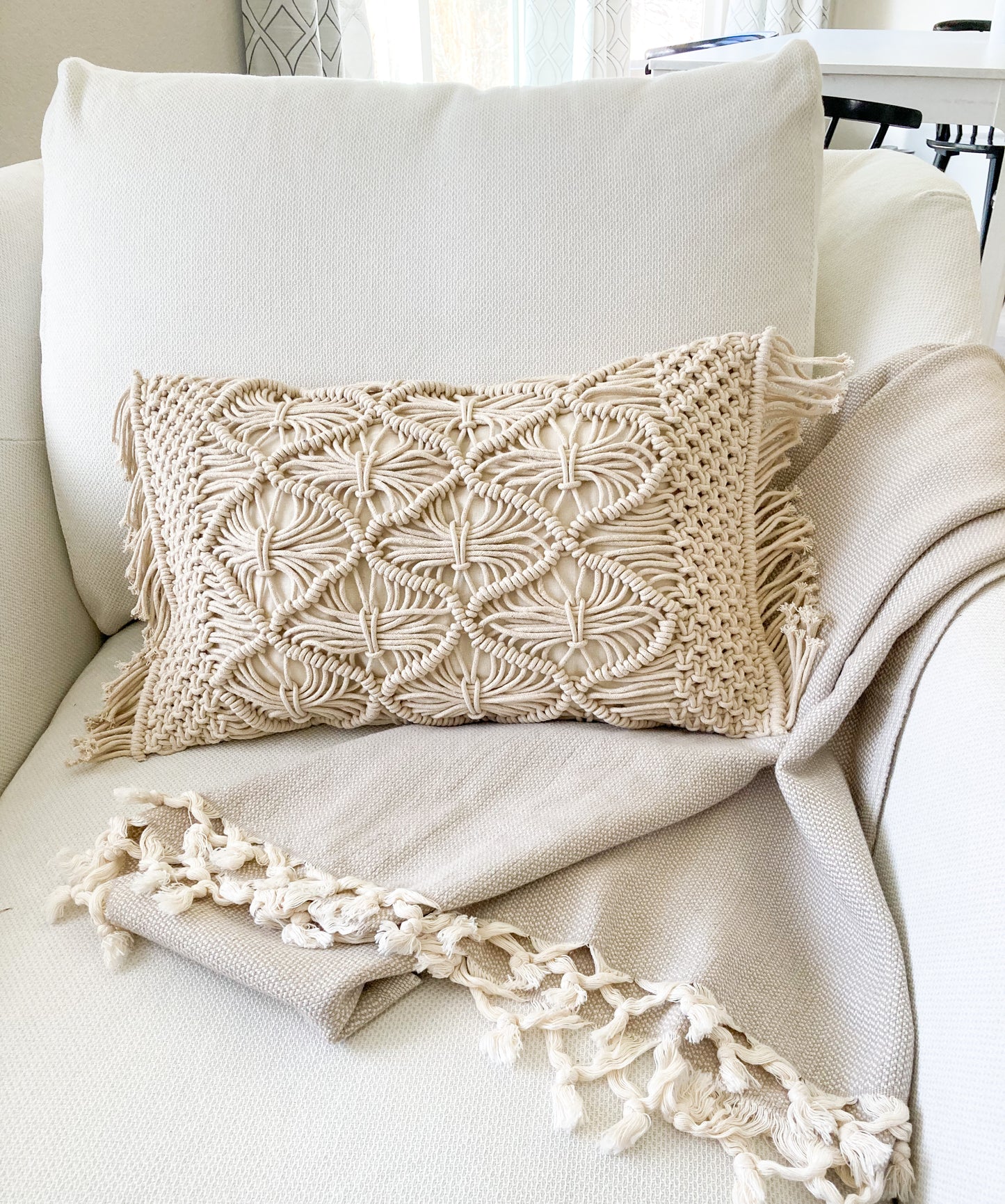 ivory lumbar macrame pillow with fringe on the side sitting on a large arm chair with a beige blanket