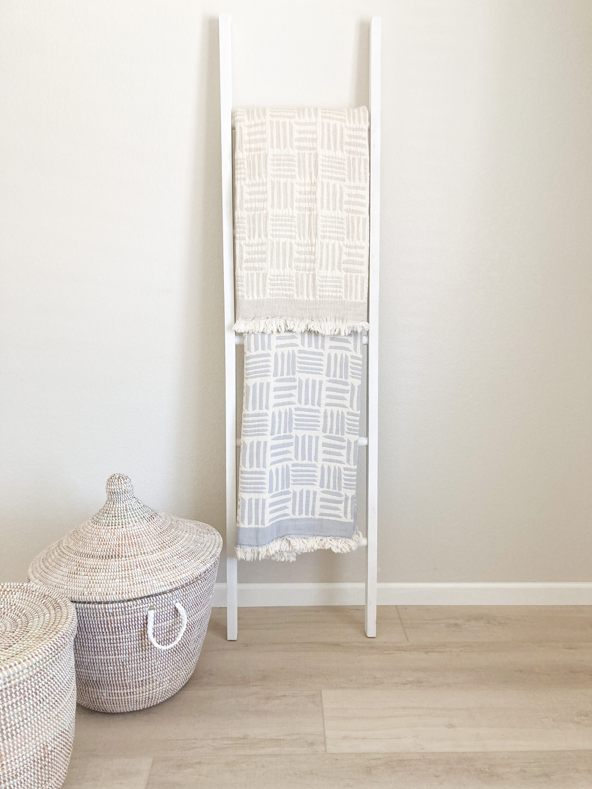 2 blankets hanging on a blanket ladder.  There top blanket is beige and ivory.  The bottom blanket is light gray and ivory. Both blankets have a modern geometric pattern.  There are 2 baskets with lids to the bottom left of the ladder.