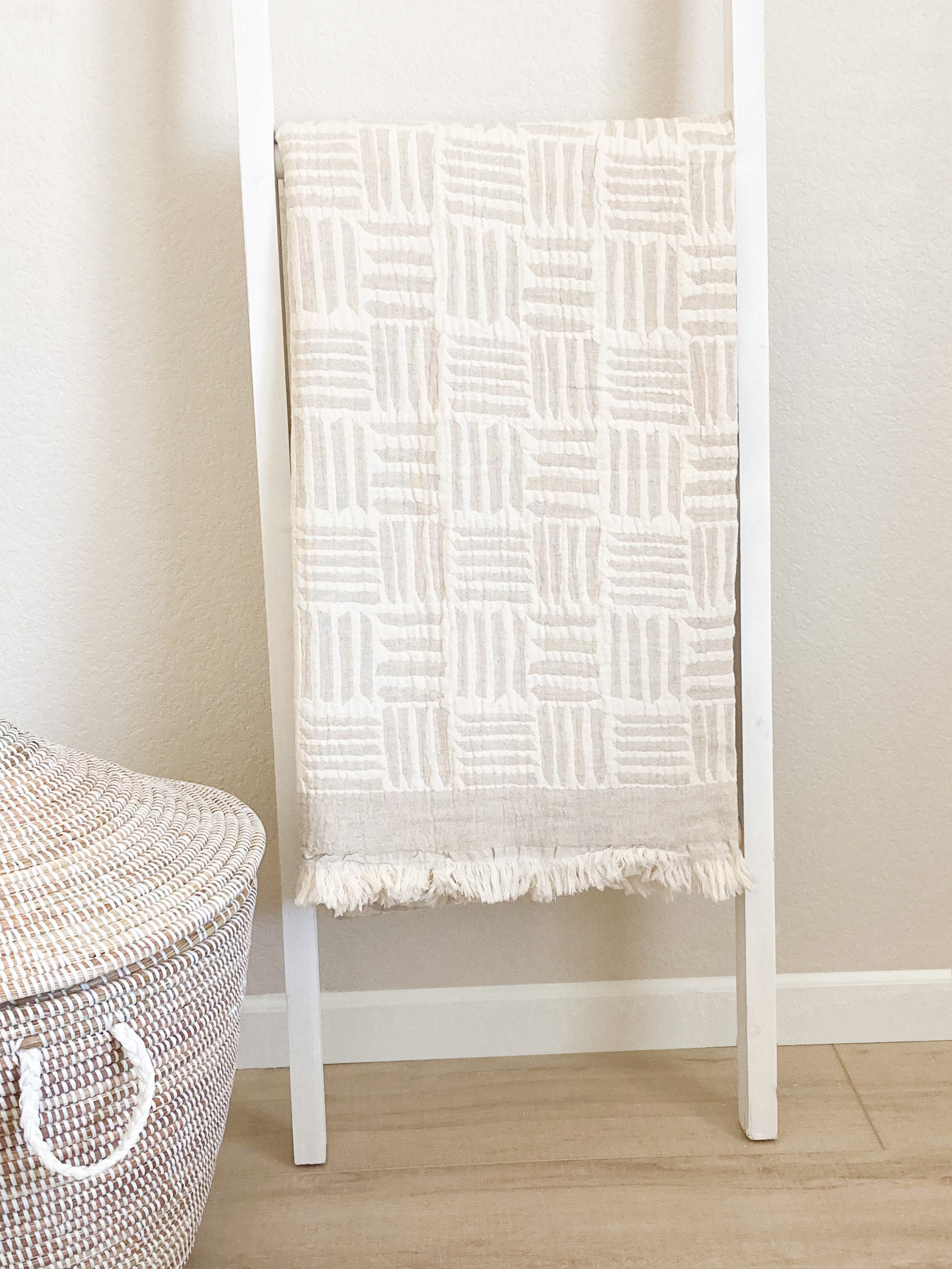 ivory and beige blanket hanging on a ladder. The blanket has a modern geometric pattern. There is a large basket with the lid to the  bottom left of the blanket ladder.