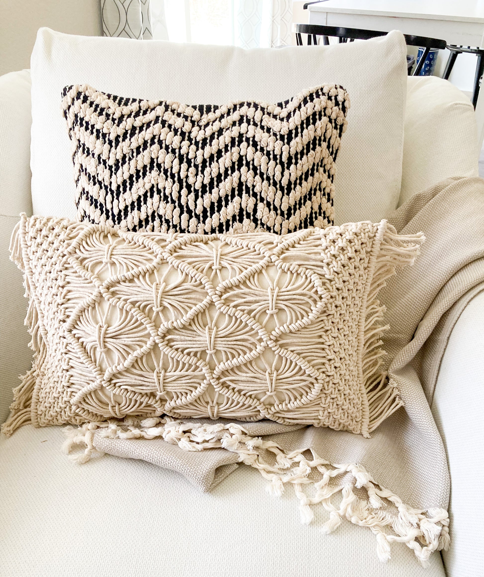2 pillows on an ivory arm chair with a beige blanket. Pillow in the front is an ivory lumbar macrame pillow with fringe on 2 sides.  The pillow in the back is an 18inch square with a zig zag pattern in ivory and dark gray